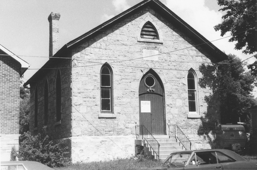 A limestone church with a gabled roof, a gothic gable vent, and two lancet windows on either side of a pointed arch front door reached by a set of stairs.